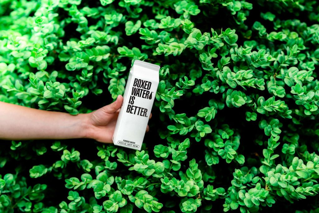 A hand holding a carton of water in front of a bush